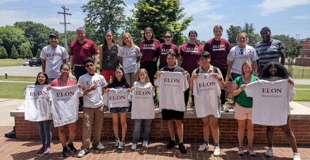 A group of students and faculty pose outdoors in two rows, smiling and holding gray T-shirts that read “Elon Biomechanics.”